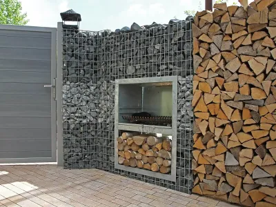 Ein langlebiger Garten-Grill verbaut in einer Gabionenwand aus Naturstein direkt neben einem modernen Zaun.