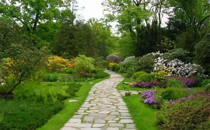 Ein geschwungener Gartenweg aus hellem Pflaster eingebettet in eine blühende Parkanlage mit Bäumen und Sträuchern.