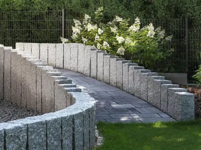 Vertikale Palisaden aus Naturstein säumen eine sanft geschwungene Rampe in einer grünen Gartenanlage.