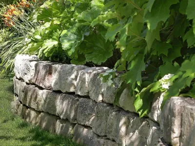 Eine solide Trockenmauer aus Naturstein stützt ein üppig bepflanztes Beet entlang der Rasenfläche.