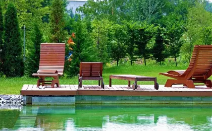 Stilvolles Holzdeck mit hölzernen Liegestühlen an einem klaren Schwimmteich. Moderne Teiche für Freizeit und Garten.