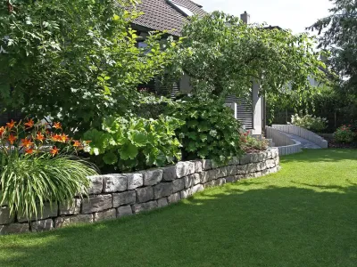 Eine Trockenmauer aus Naturstein grenzt ein üppiges Beet von saftigem Rollrasen ab.