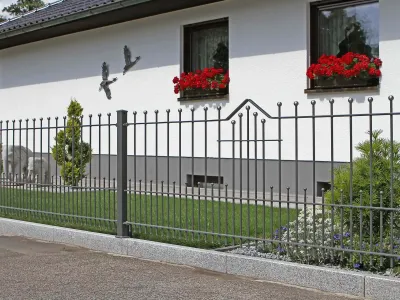 Stilvoller Metallzaun auf einem Sockel aus Naturstein vor einer weißen Hausfassade mit Blumen.