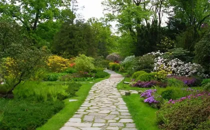 Ein geschwungener Gartenweg aus hellem Pflaster eingebettet in eine blühende Parkanlage mit Bäumen und Sträuchern.
