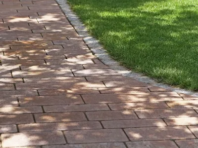 Ein gartengestalteter Fußweg aus rötlichen Pflastersteinen mit einer sauberen Einfassung aus Naturstein.